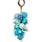 A cluster of blue and turquoise beaded charms hangs from a gold-colored clasp. The beads vary in size, texture, and shade, including some with glitter and gold accents. The keychain is suspended against a white background with black dots.
