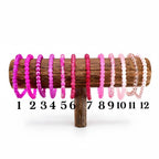 A wooden display stand holds twelve beaded bracelets in shades of pink and purple. The bracelets are numbered 1 through 12. The beads vary in size and texture, with some appearing smooth and others more textured.