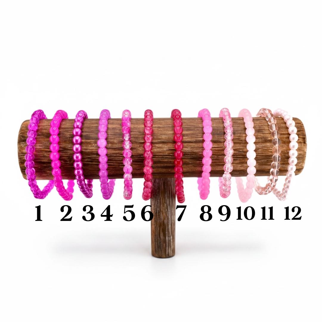 A wooden display stand holds twelve beaded bracelets in shades of pink and purple. The bracelets are numbered 1 through 12. The beads vary in size and texture, with some appearing smooth and others more textured.