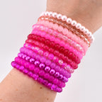 A collection of beaded bracelets in shades of pink and magenta. The bracelets vary in style, including pearl, clear, and solid beads. The bracelets are stacked on a wrist, showcasing the range of colors and textures.