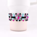 A stack of beaded bracelets in various colors, including black, pink, blue, and silver. The bracelets are wrapped around a white mug. The beads are small and round, with some featuring striped patterns.