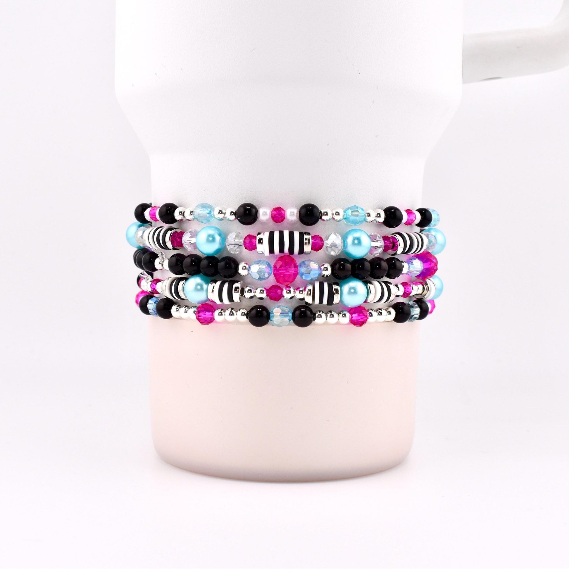 A stack of beaded bracelets in various colors, including black, pink, blue, and silver. The bracelets are wrapped around a white mug. The beads are small and round, with some featuring striped patterns.