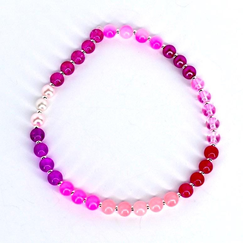 A beaded bracelet featuring a gradient of pink and red hues. The bracelet is composed of round beads in shades of fuchsia, magenta, rose, and red, separated by small silver-colored spacers. The beads vary in size and material, creating a visually appealing accessory.