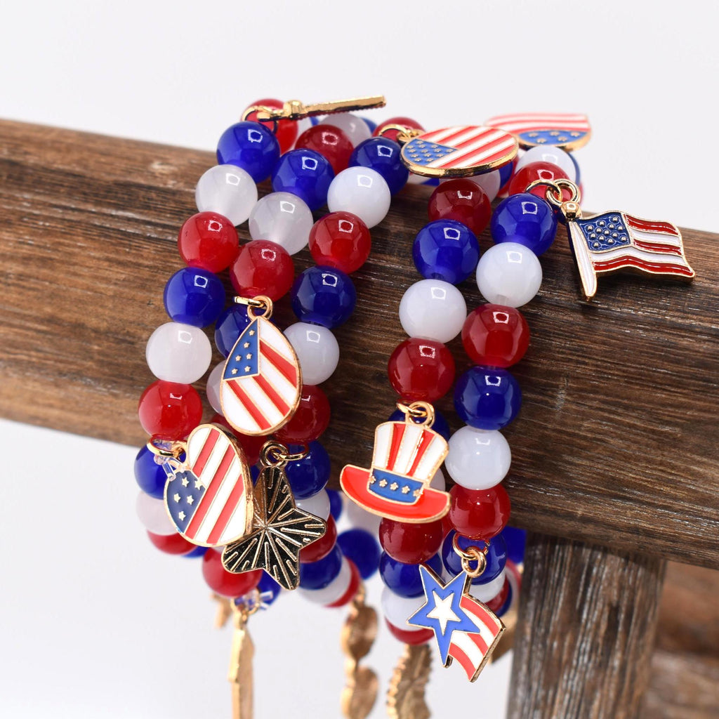 Beaded bracelets in red, white, and blue, adorned with patriotic charms. The bracelets feature round beads and charms shaped like the American flag, a top hat, and a star. The bracelets are displayed on a wooden surface.