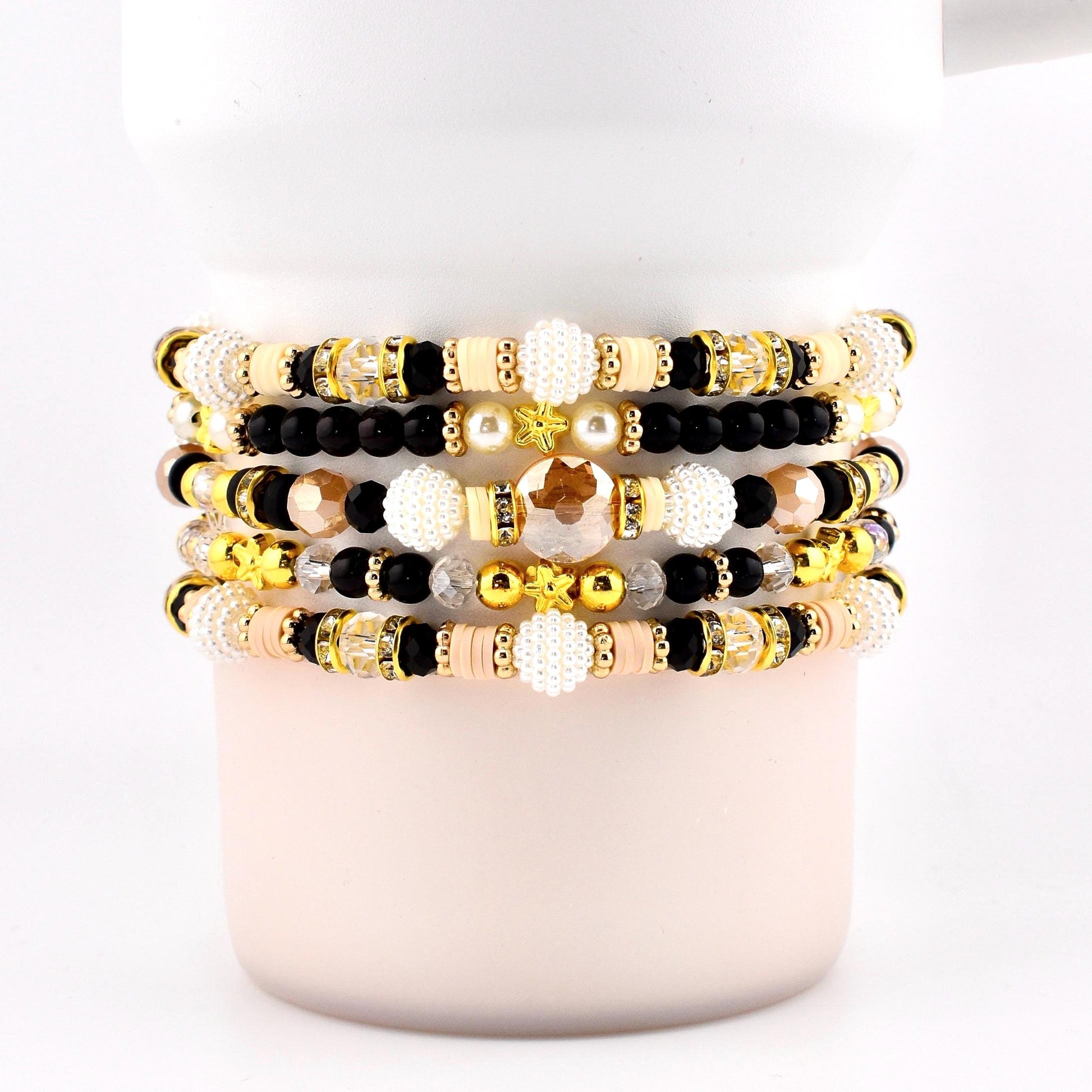Stack of beaded bracelets in various colors, including gold, black, cream, and peach. The bracelets feature a mix of beads, including round, faceted, and pearl-like beads. Some bracelets have gold accents and star-shaped beads.