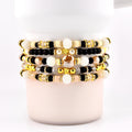 Stack of beaded bracelets in various colors, including gold, black, cream, and peach. The bracelets feature a mix of beads, including round, faceted, and pearl-like beads. Some bracelets have gold accents and star-shaped beads.