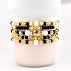 Stack of beaded bracelets in various colors, including gold, black, cream, and peach. The bracelets feature a mix of beads, including round, faceted, and pearl-like beads. Some bracelets have gold accents and star-shaped beads.