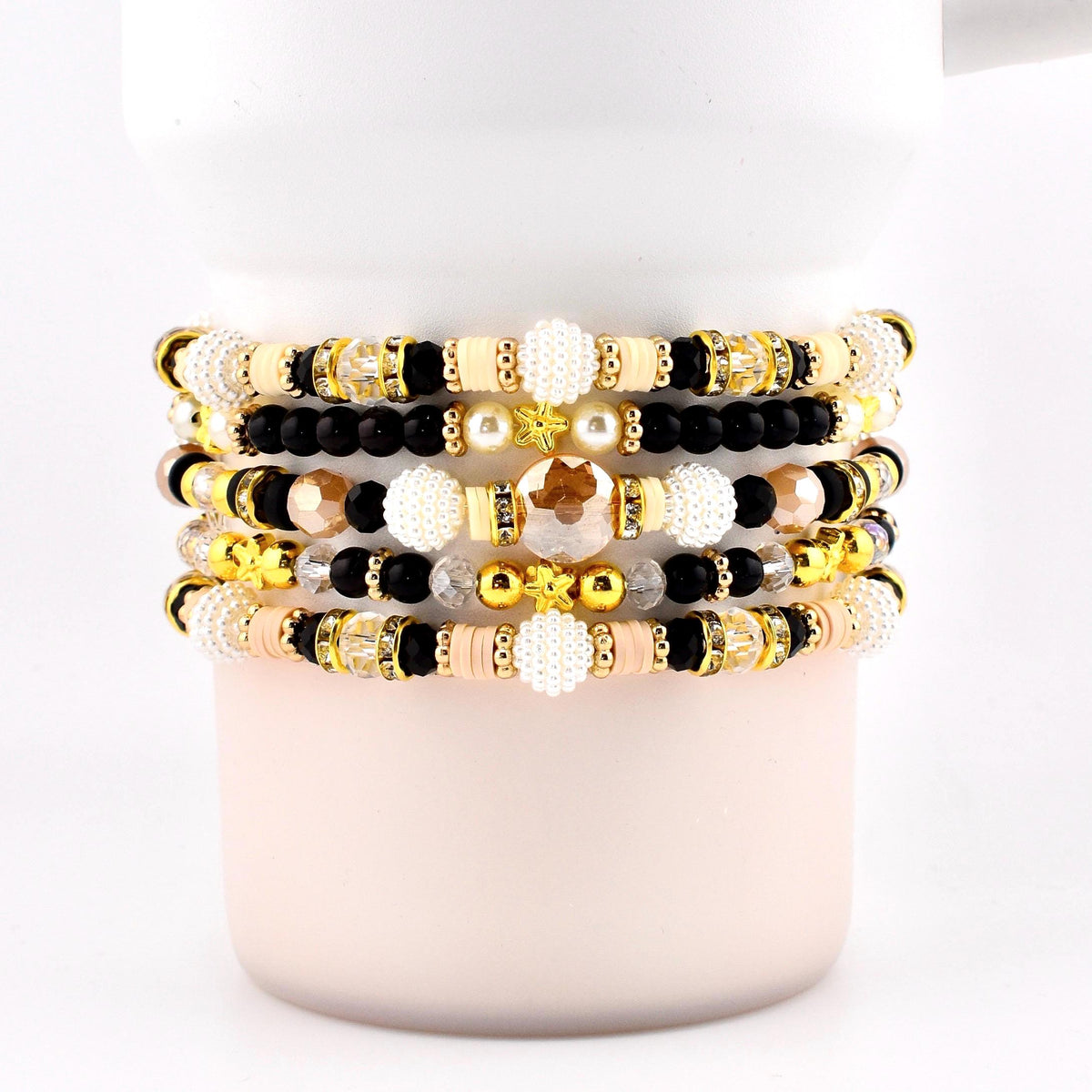 Stack of beaded bracelets in various colors, including gold, black, cream, and peach. The bracelets feature a mix of beads, including round, faceted, and pearl-like beads. Some bracelets have gold accents and star-shaped beads.