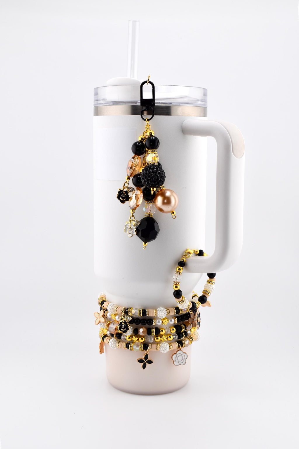 White Stanley tumbler with a black and gold beaded charm and several beaded bracelets. The charm features black, gold, and peach-colored beads. The bracelets have gold, black, and white beads with flower and cross charms.