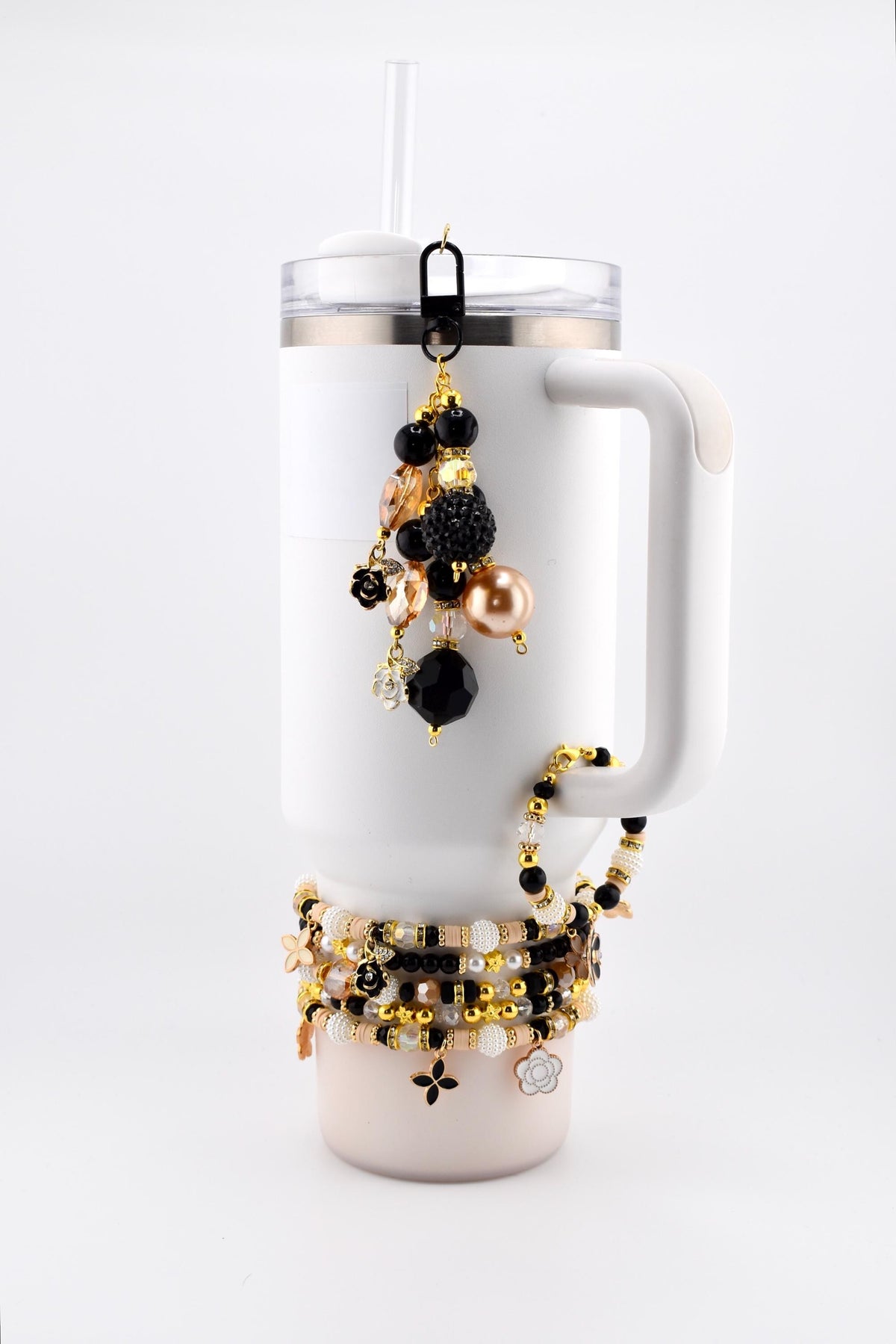White Stanley tumbler with a black and gold beaded charm and several beaded bracelets. The charm features black, gold, and peach-colored beads. The bracelets have gold, black, and white beads with flower and cross charms.