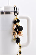 A beaded straw charm with a black clasp, attached to a white mug. The charm features a variety of beads in black, gold, and champagne colors, including faceted, round, and floral designs. The charm is a decorative accessory.