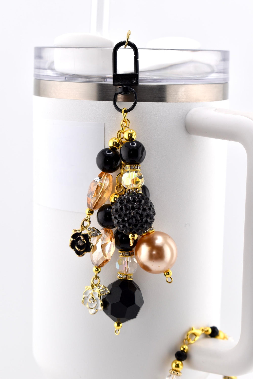 A decorative charm for a tumbler or mug, featuring a black clip and a dangling assortment of beads. The beads include black, gold, and clear faceted beads, a large gold pearl, and small gold and white flower charms.