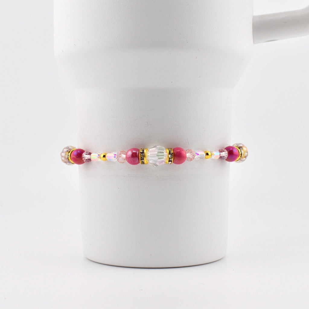 A beaded bracelet with pink, gold, and clear beads. The bracelet features a central cluster of larger beads, including a clear, faceted bead and several pink beads. The bracelet is strung on a thin, flexible cord.