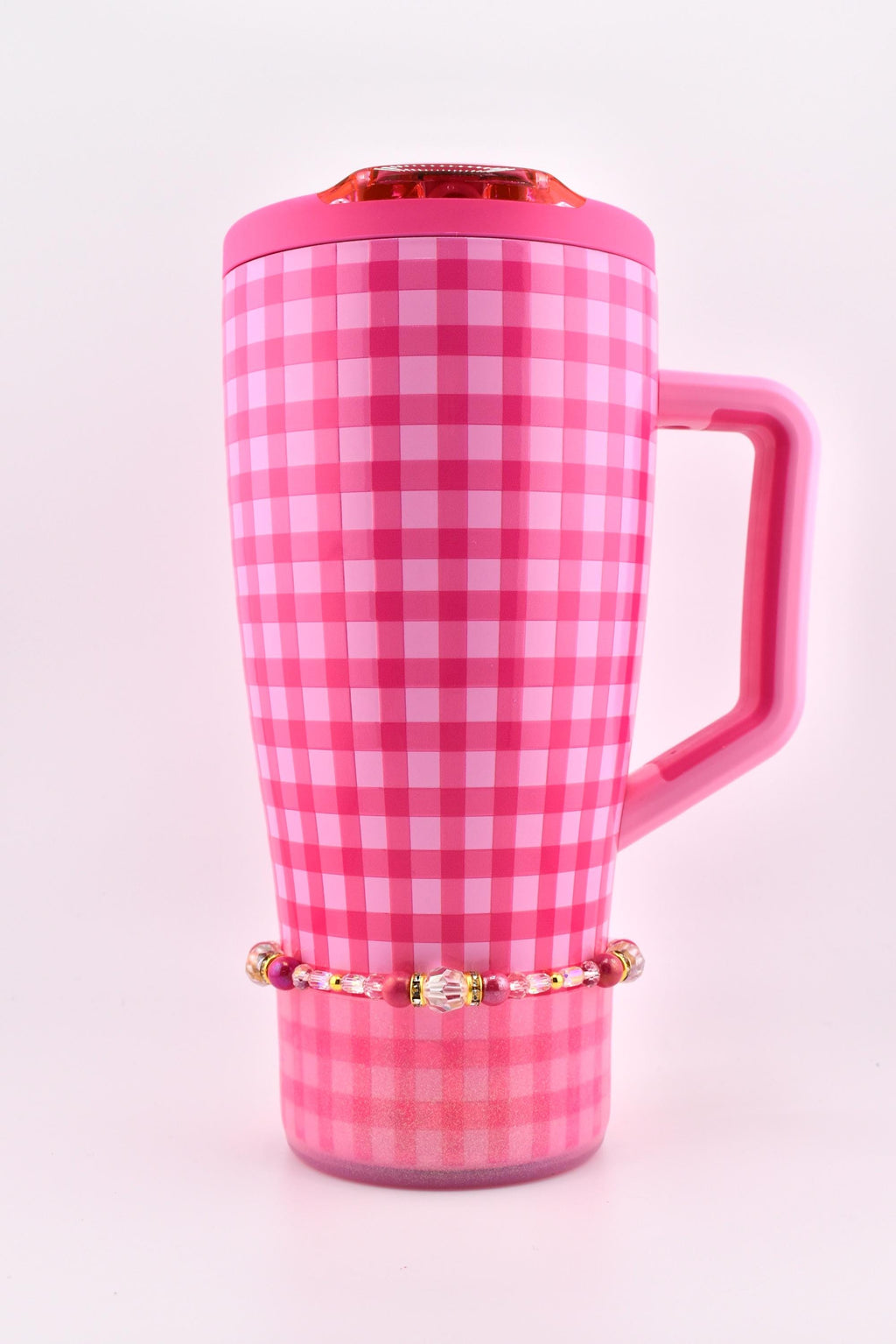 A pink and white checkered travel mug with a matching handle and lid. A beaded bracelet with pink and gold accents is wrapped around the base of the mug. The mug has a shiny finish.
