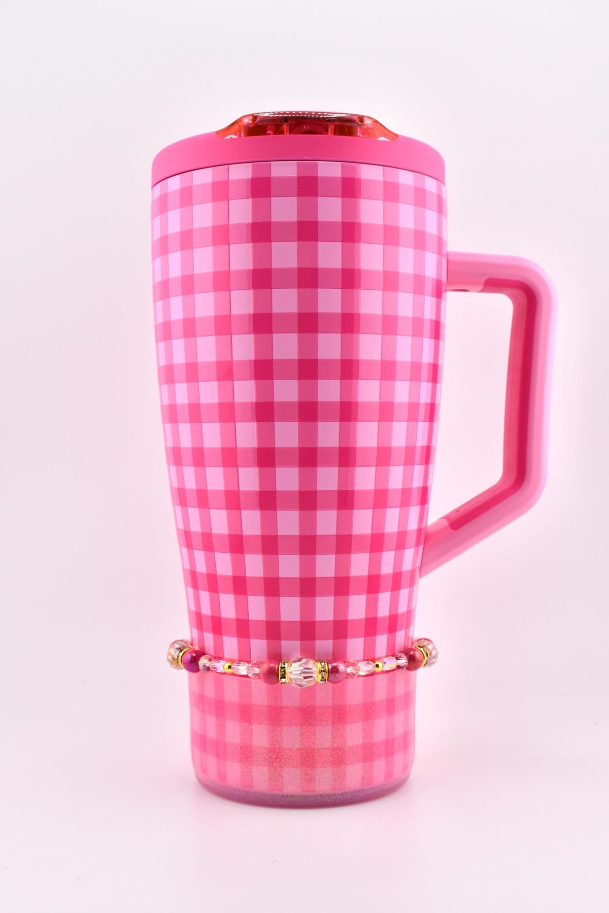A pink and white checkered travel mug with a matching handle and lid. A beaded bracelet with pink and gold accents is wrapped around the base of the mug. The mug has a shiny finish.