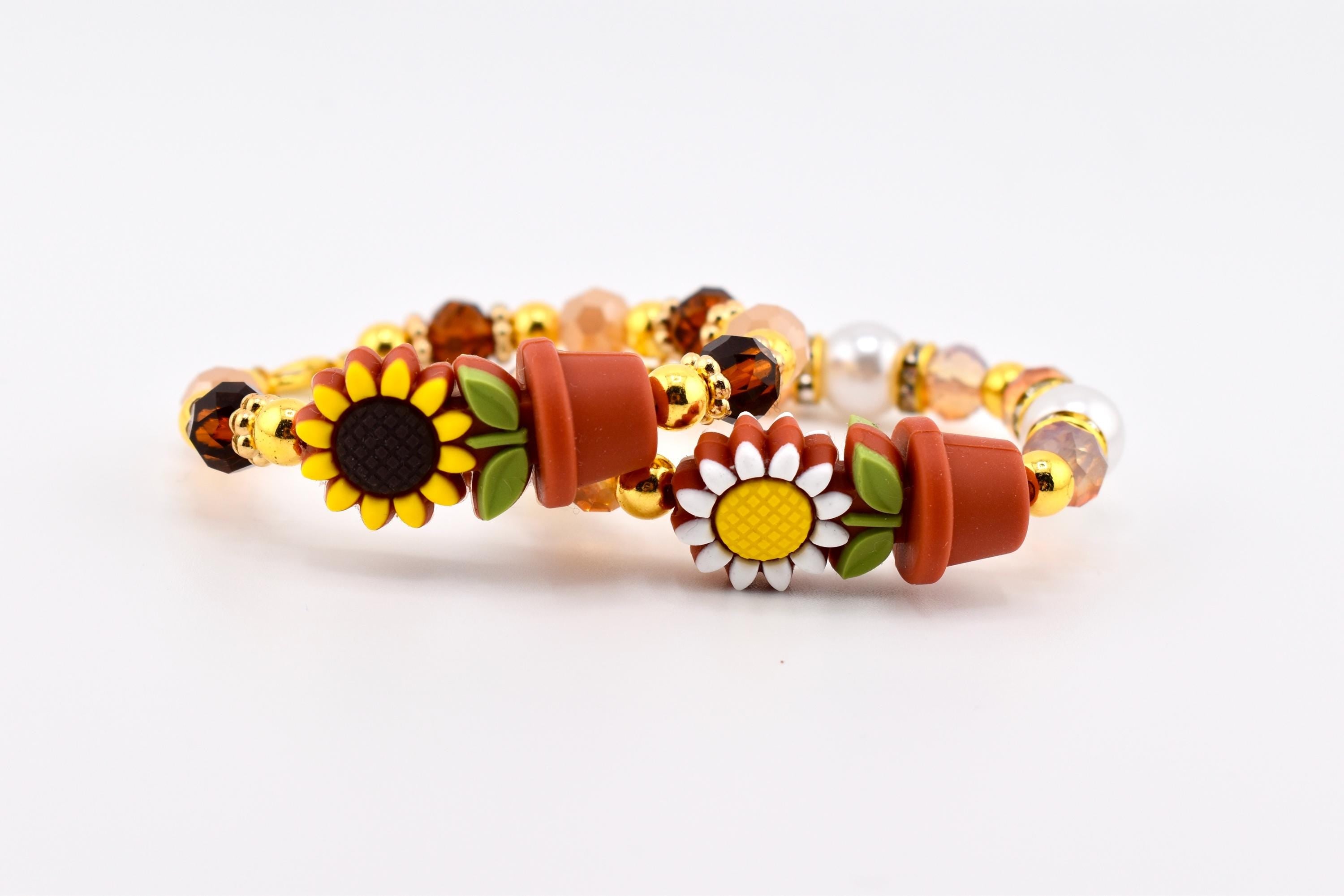 Two beaded bracelets with sunflower and flower pot charms. The bracelets feature a mix of gold, brown, and peach-colored beads, with a few white pearl beads. The sunflower charms have yellow petals and dark brown centers.