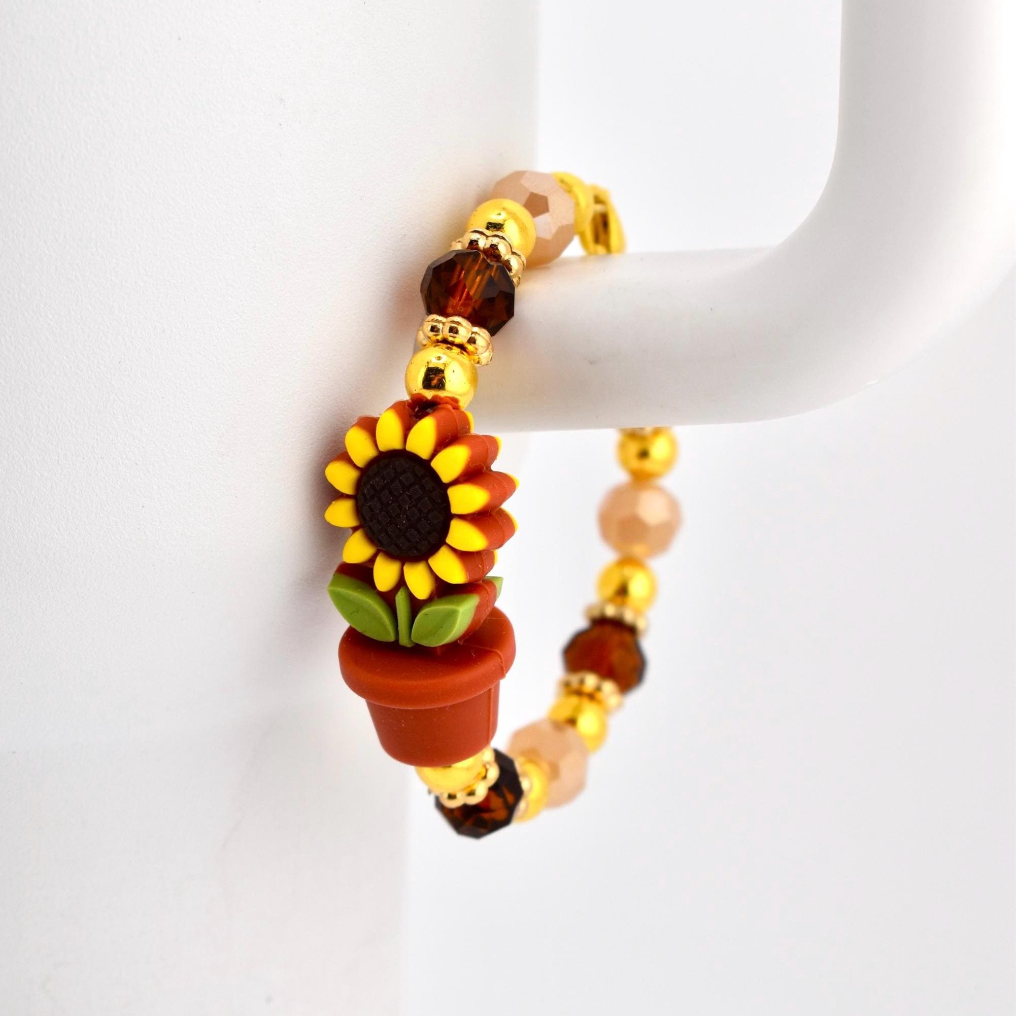 A beaded tumbler handle bracelet featuring a sunflower in a terracotta pot. The bracelet has gold-colored accents and is strung with brown, peach, and gold beads. The sunflower has yellow petals and a dark brown center.