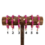 Six pink beaded bracelets with gold accents and charms, displayed on a wooden stand. Each bracelet features a different charm, including a flower, heart, bow, and seashell. The bracelets are numbered 1 through 6.