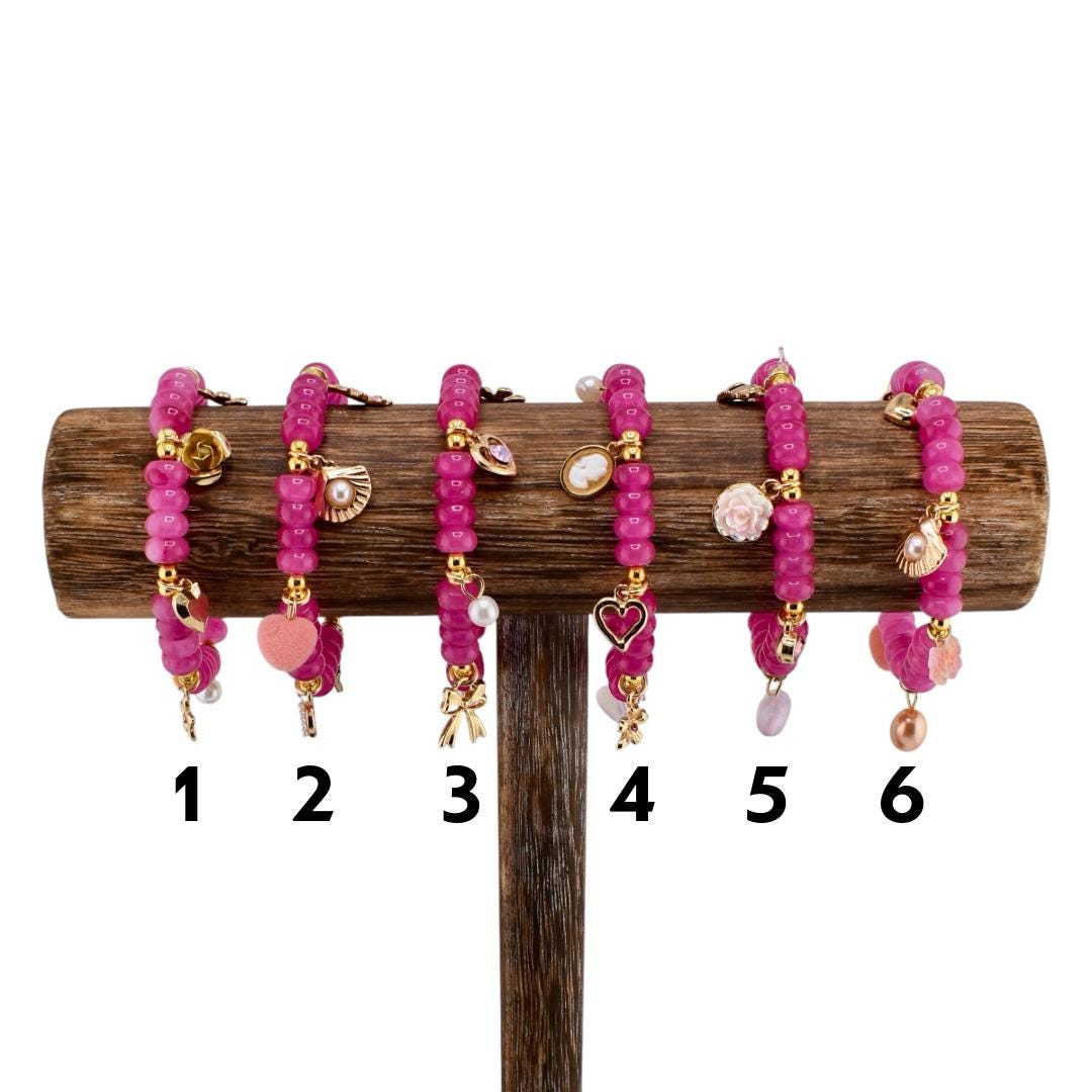 Six pink beaded bracelets with gold accents and charms, displayed on a wooden stand. Each bracelet features a different charm, including a flower, heart, bow, and seashell. The bracelets are numbered 1 through 6.