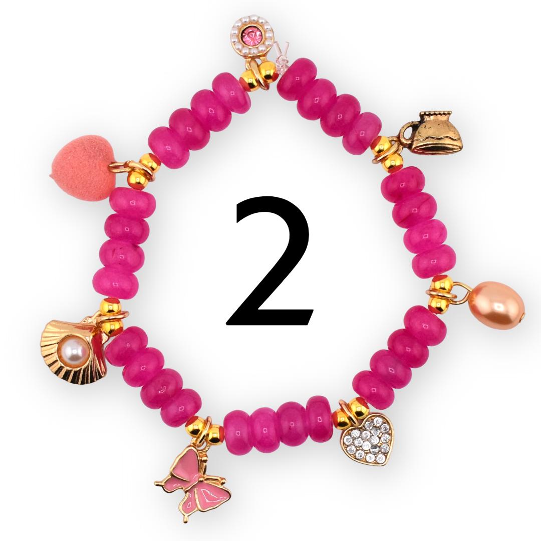 A magenta beaded bracelet with gold accents and various charms, including a heart, butterfly, seashell, and a small teapot. The bracelet also features a pink heart-shaped charm and a pearl-shaped charm. The number "2" is in the center.