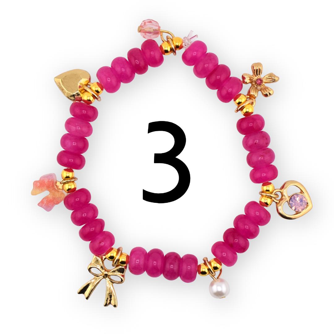 A magenta beaded bracelet with gold-colored charms. The charms include a heart, a flower, a bow, and a pearl. The number "3" is in the center of the bracelet.