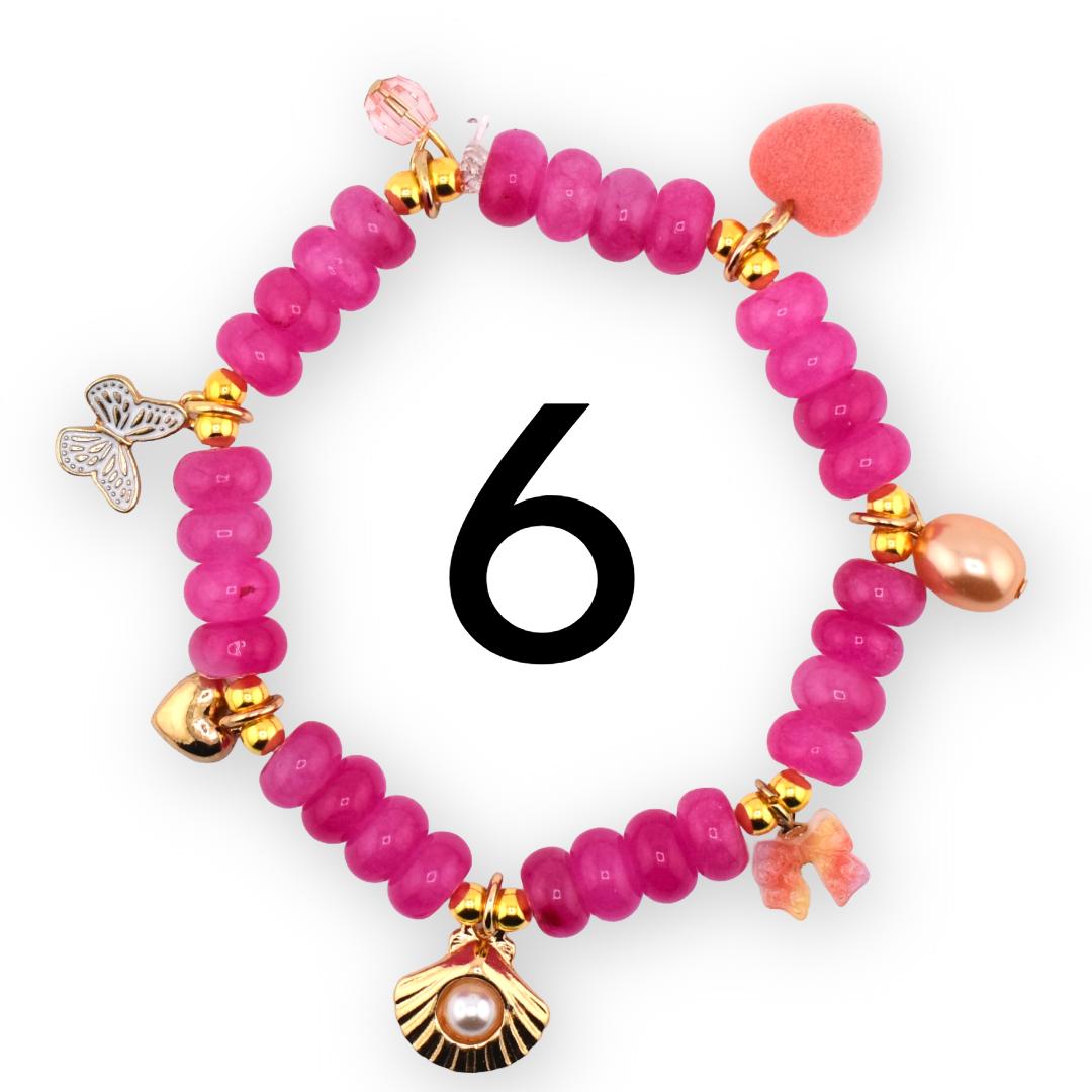 A pink beaded bracelet with gold accents and decorative charms. Charms include a butterfly, a seashell with a pearl, a heart, and a bow. The number 6 is in the center of the bracelet.