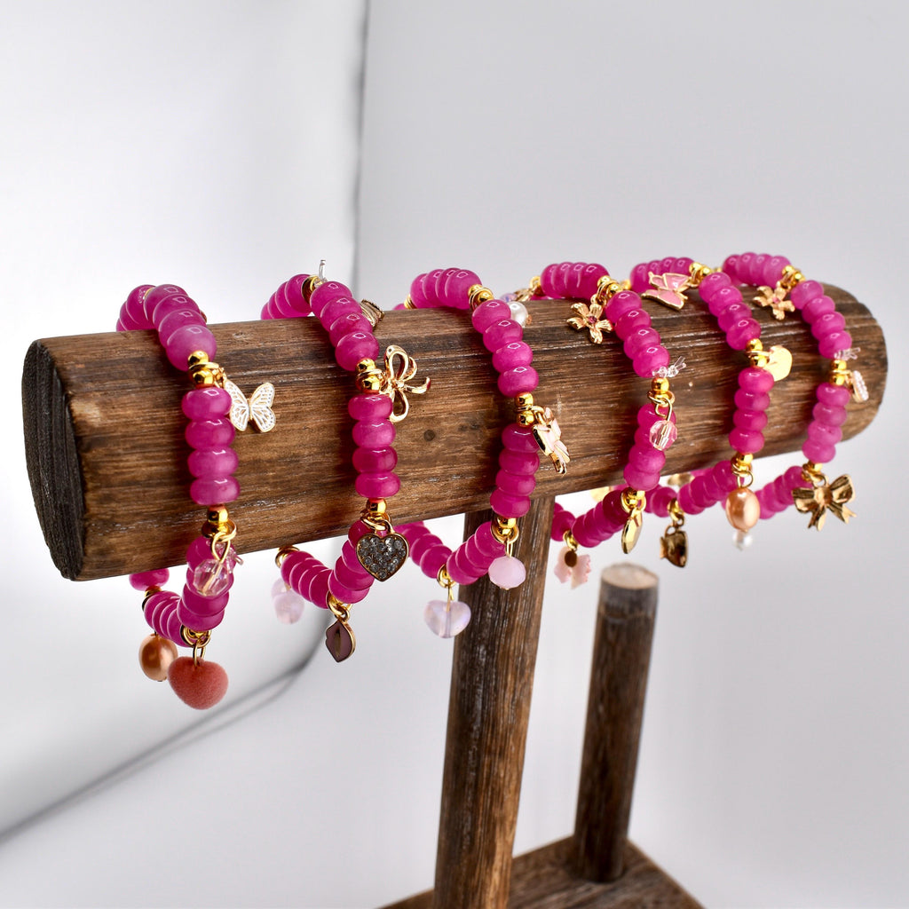 Six magenta beaded bracelets with gold accents and charms, including butterflies, bows, and hearts, are displayed on a wooden stand. The bracelets are made of round beads and have a delicate, feminine design.