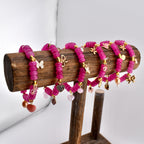 Six magenta beaded bracelets with gold accents and charms, including butterflies, bows, and hearts, are displayed on a wooden stand. The bracelets are made of round beads and have a delicate, feminine design.