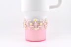 A white tumbler with a pink base is adorned with several beaded bracelets. The bracelets feature pink beads, small daisy accents, and silver-colored spacers. The bracelets are wrapped around the tumbler.