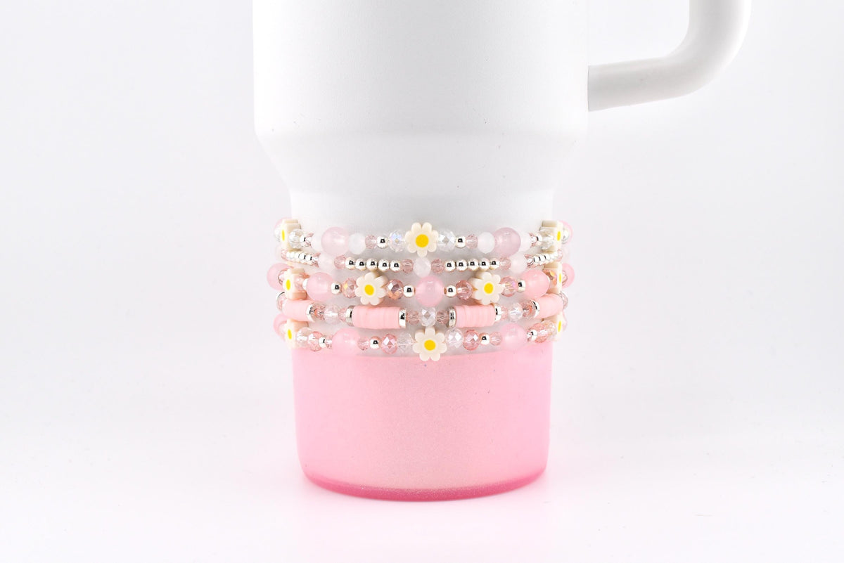 A white tumbler with a pink base is adorned with several beaded bracelets. The bracelets feature pink beads, small daisy accents, and silver-colored spacers. The bracelets are wrapped around the tumbler.