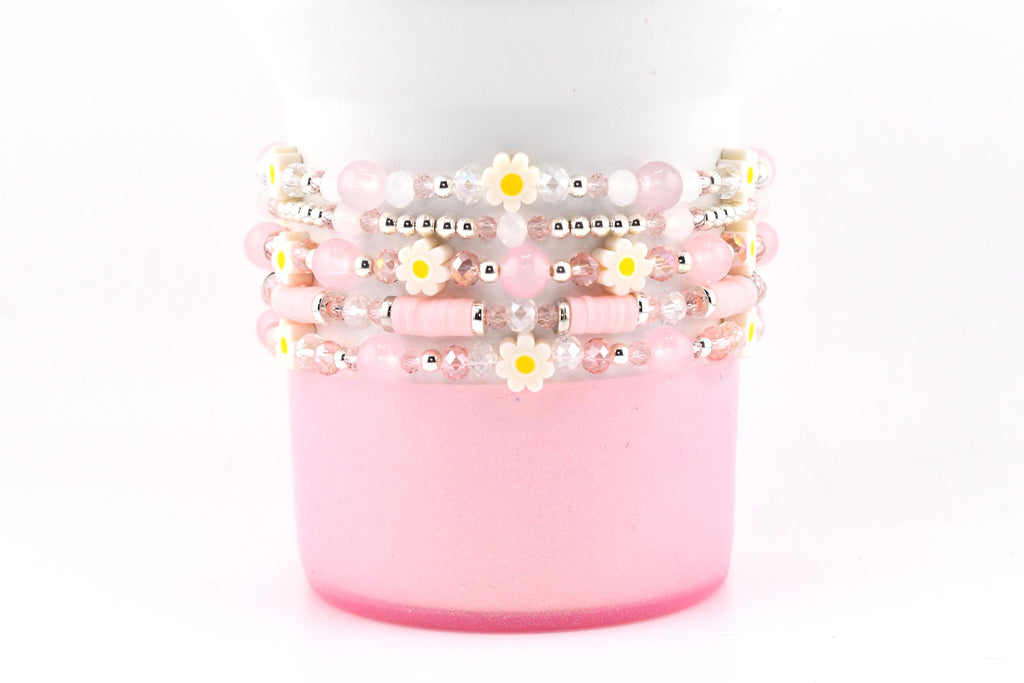 Stack of beaded bracelets in shades of pink, white, and silver. The bracelets feature small daisy accents with yellow centers. The bracelets are stacked on a pink cylindrical object against a white background.