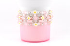 Stack of beaded bracelets in shades of pink, white, and silver. The bracelets feature small daisy accents with yellow centers. The bracelets are stacked on a pink cylindrical object against a white background.