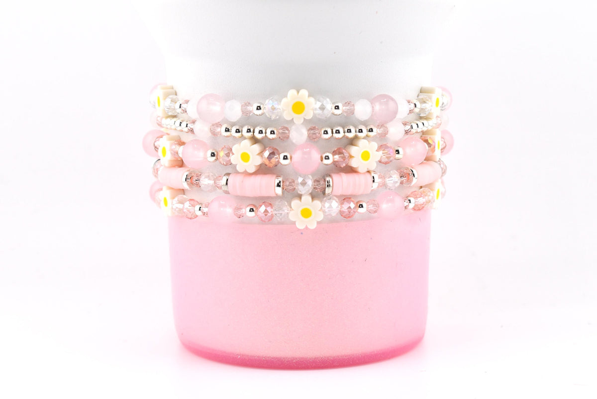 Stack of beaded bracelets in shades of pink, white, and silver. The bracelets feature small daisy accents with yellow centers. The bracelets are stacked on a pink cylindrical object against a white background.