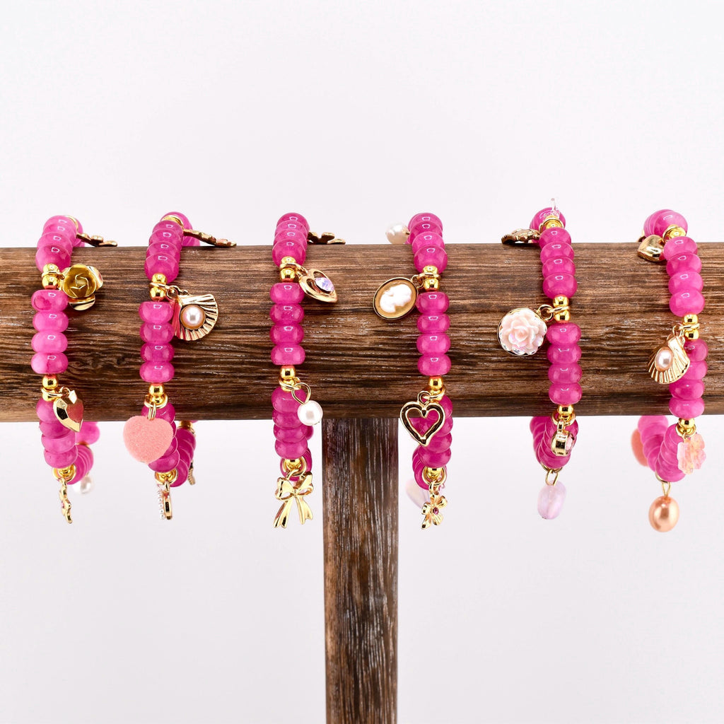 A collection of pink beaded bracelets with gold-colored charms. Each bracelet features a different charm, including a heart, a bow, a pearl, a seashell, and a flower. The bracelets are displayed on a wooden stand.