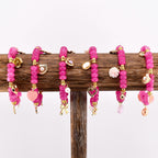 A collection of pink beaded bracelets with gold-colored charms. Each bracelet features a different charm, including a heart, a bow, a pearl, a seashell, and a flower. The bracelets are displayed on a wooden stand.