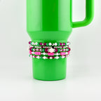 A stack of beaded bracelets in shades of pink, white, green, and silver are wrapped around a bright green tumbler. The bracelets feature various bead sizes and textures, including round and cylindrical beads, creating a layered effect.
