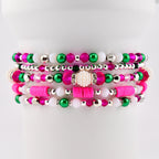 Stack of beaded bracelets in shades of pink, green, white, and silver. The bracelets feature various bead sizes and shapes, including round, disc, and pearl-like beads. The bracelets are stacked on a white surface.