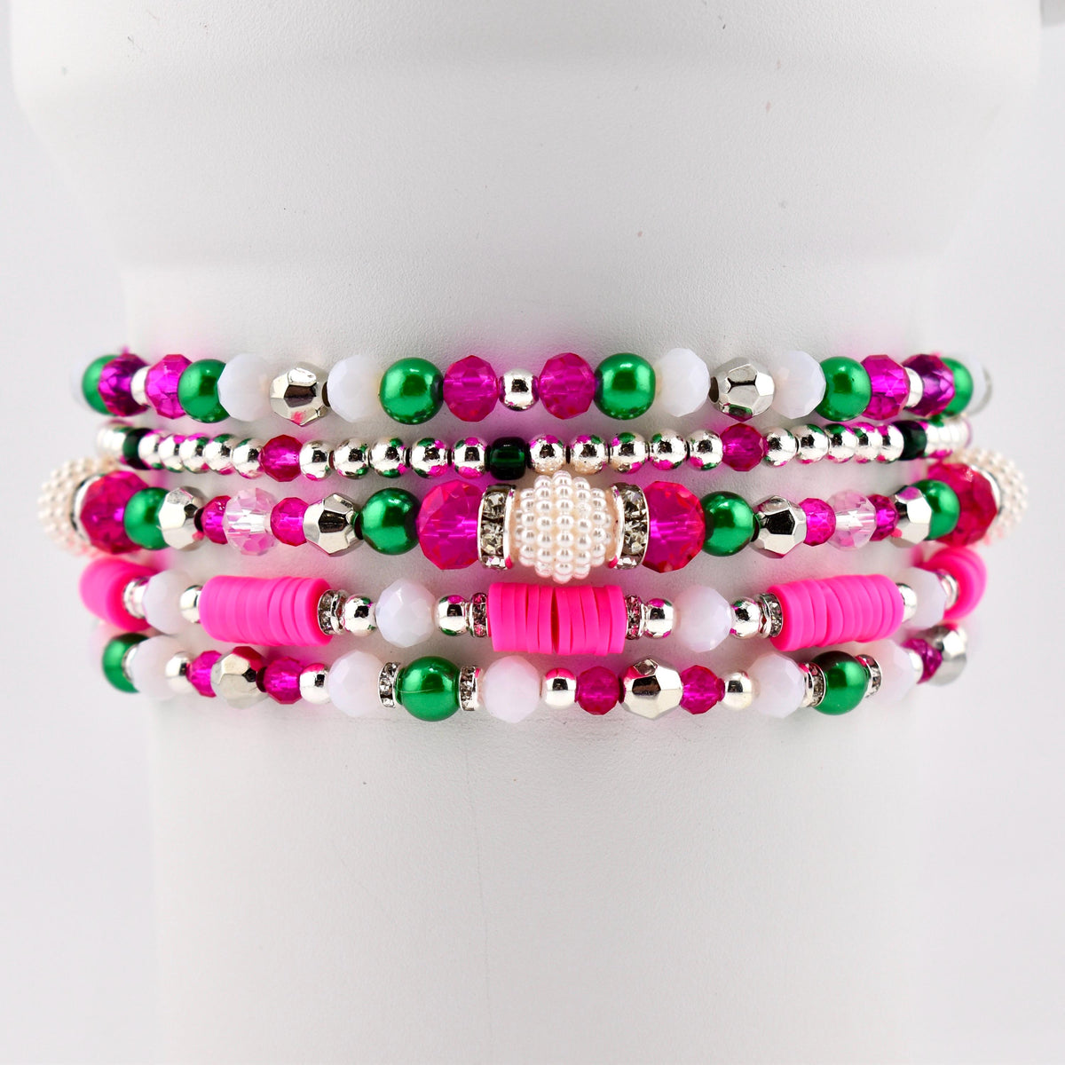 Stack of beaded bracelets in shades of pink, green, white, and silver. The bracelets feature various bead sizes and shapes, including round, disc, and pearl-like beads. The bracelets are stacked on a white surface.