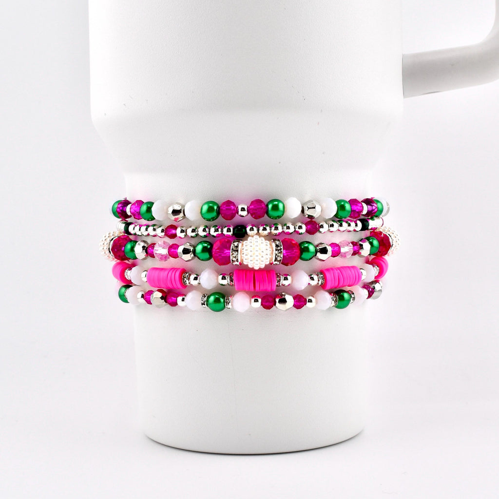 Stack of beaded bracelets in shades of pink, green, white, and silver. The bracelets are displayed on a white mug. The beads vary in size and material, creating a layered effect. The bracelets are a mix of round and flat beads.