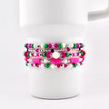 Stack of beaded bracelets in shades of pink, green, white, and silver. The bracelets are displayed on a white mug. The beads vary in size and material, creating a layered effect. The bracelets are a mix of round and flat beads.