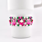 Stack of beaded bracelets in shades of pink, green, white, and silver. The bracelets are displayed on a white mug. The beads vary in size and material, creating a layered effect. The bracelets are a mix of round and flat beads.