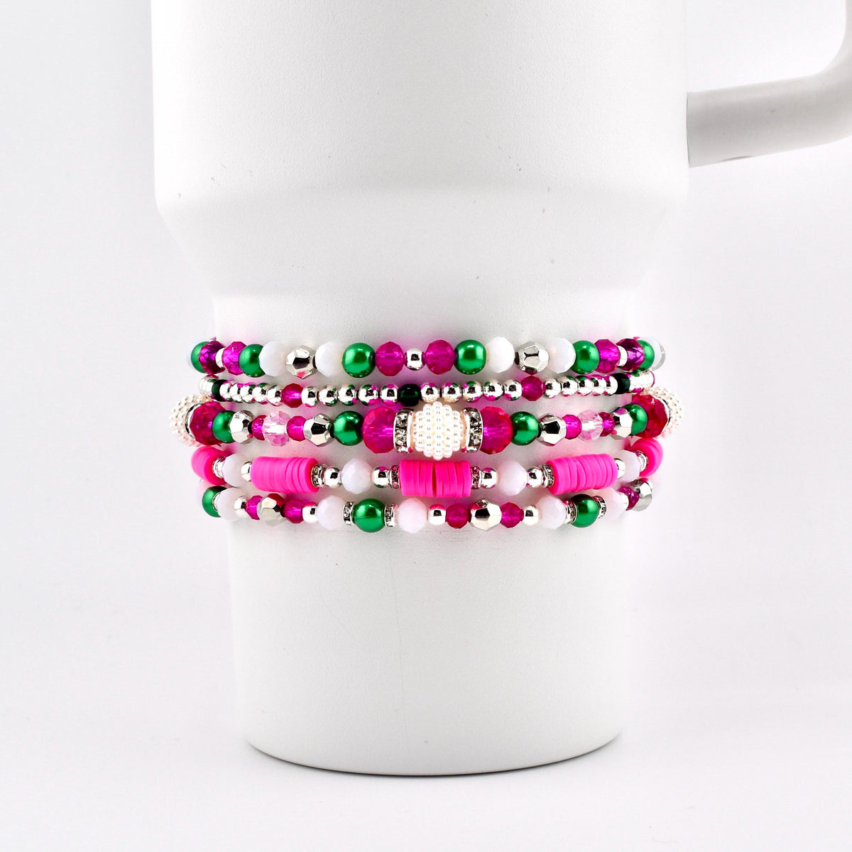 Stack of beaded bracelets in shades of pink, green, white, and silver. The bracelets are displayed on a white mug. The beads vary in size and material, creating a layered effect. The bracelets are a mix of round and flat beads.