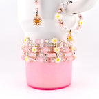 A collection of pink and white beaded bracelets, earrings, and a ring. The bracelets feature daisy-shaped beads and clear crystal beads. The earrings and ring have daisy charms. The jewelry is displayed on a pink cup against a white background.