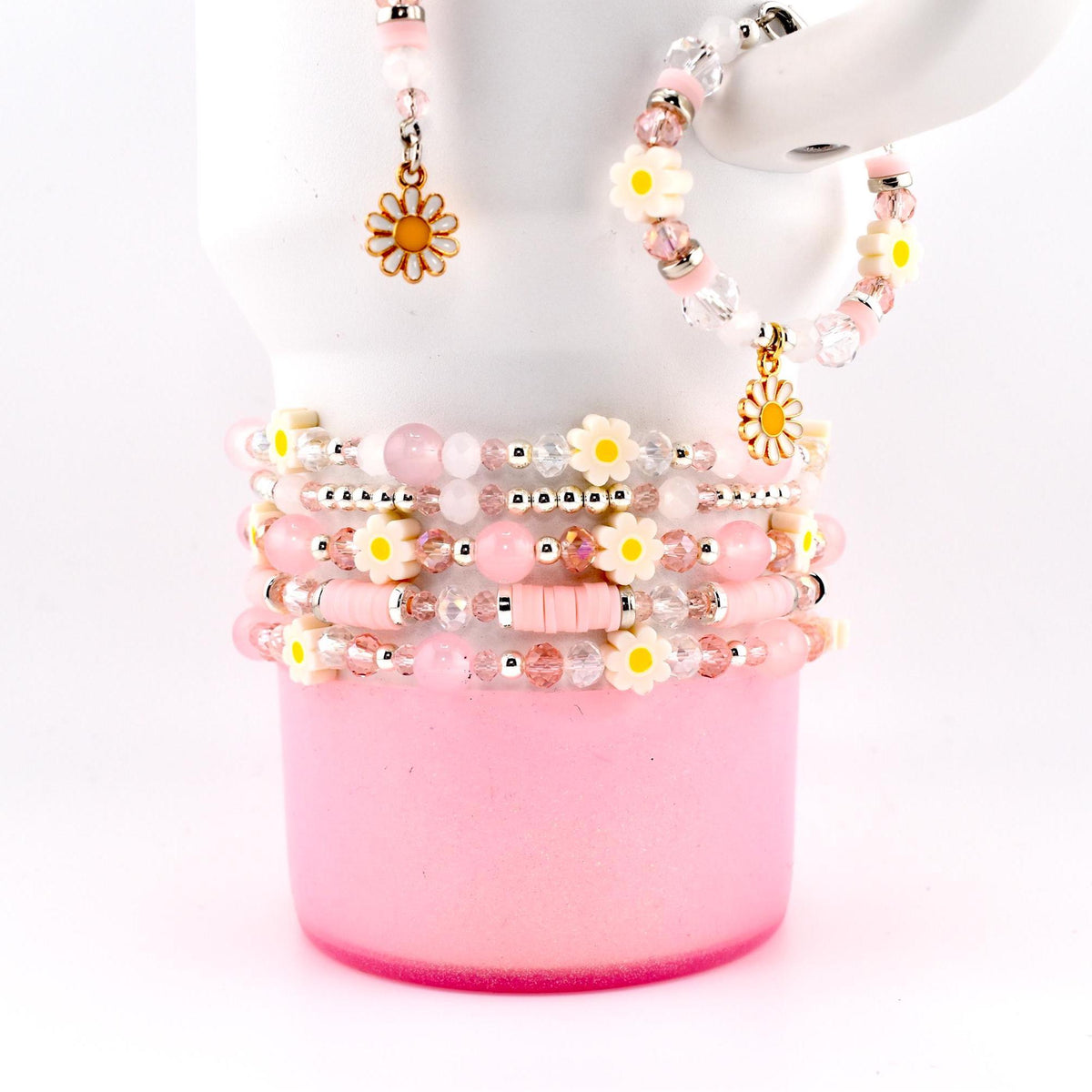 A collection of pink and white beaded bracelets, earrings, and a ring. The bracelets feature daisy-shaped beads and clear crystal beads. The earrings and ring have daisy charms. The jewelry is displayed on a pink cup against a white background.