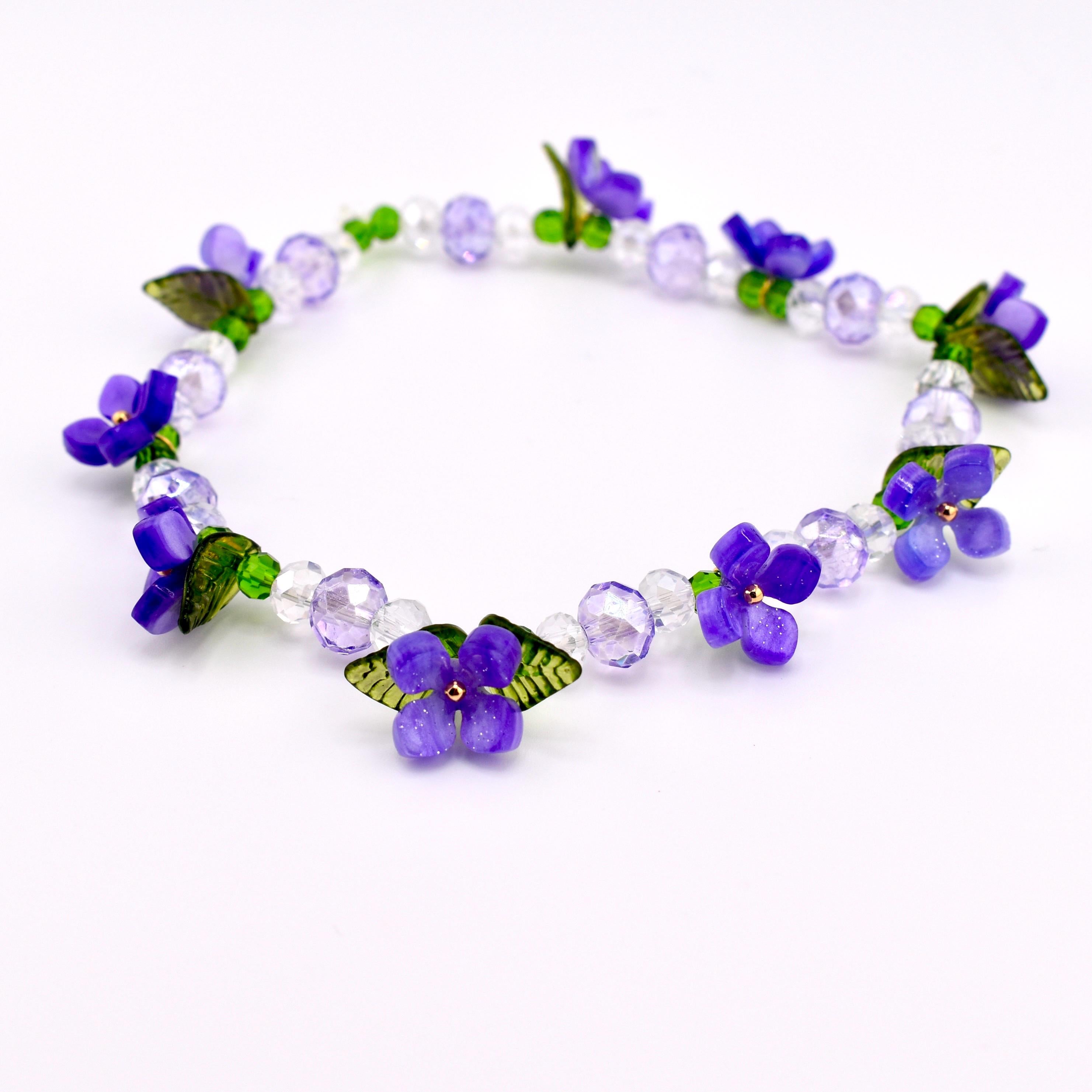 A delicate bracelet featuring vibrant purple flower charms, green leaves, and clear crystal beads. The floral design is interspersed with faceted beads and small green beads, creating a charming and elegant accessory.