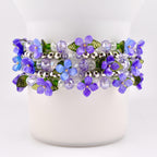 A beaded bracelet featuring a floral design with purple and light blue flowers, green leaves, and clear and silver beads. The bracelet is wrapped around a white cylindrical object.