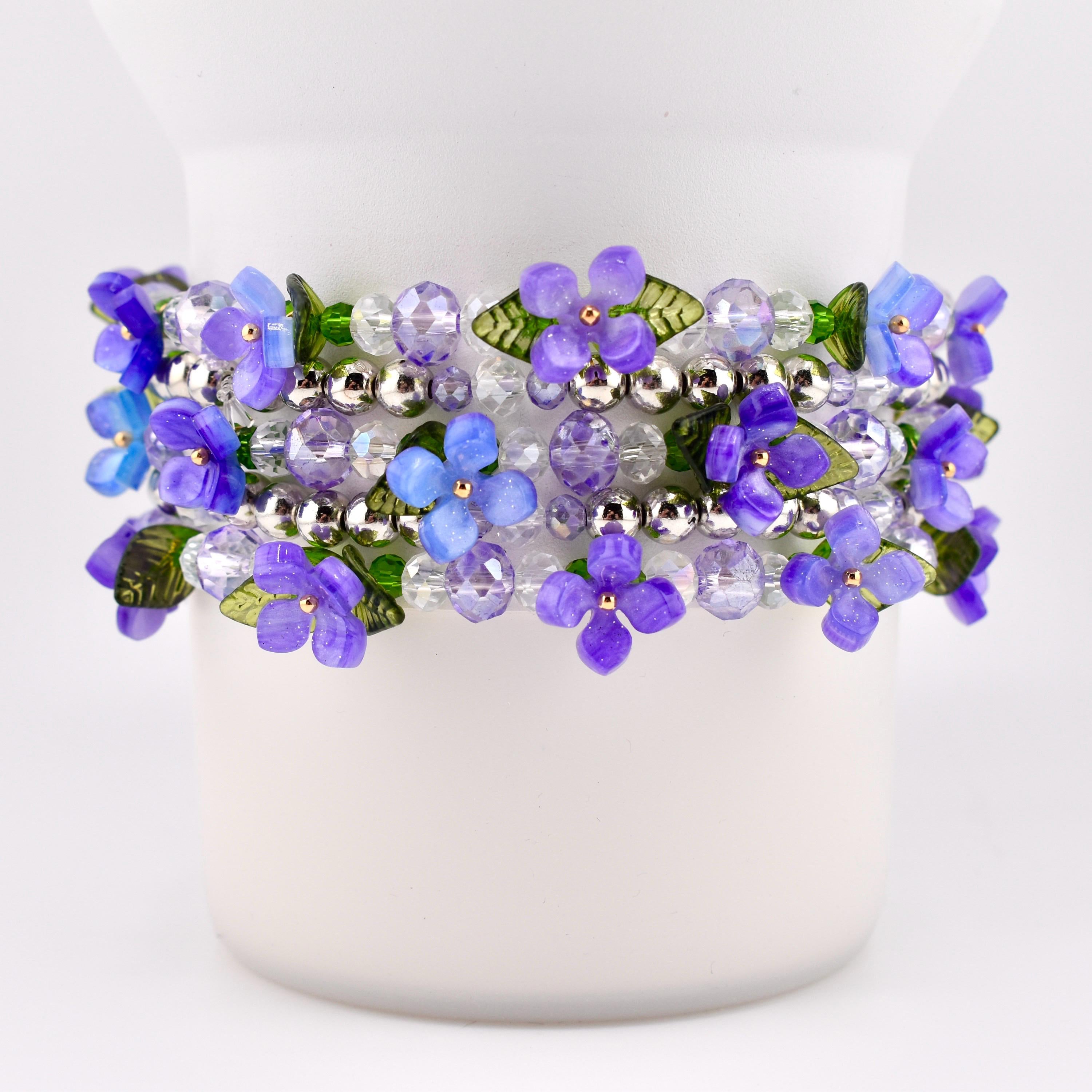 A beaded bracelet featuring a floral design with purple and light blue flowers, green leaves, and clear and silver beads. The bracelet is wrapped around a white cylindrical object.