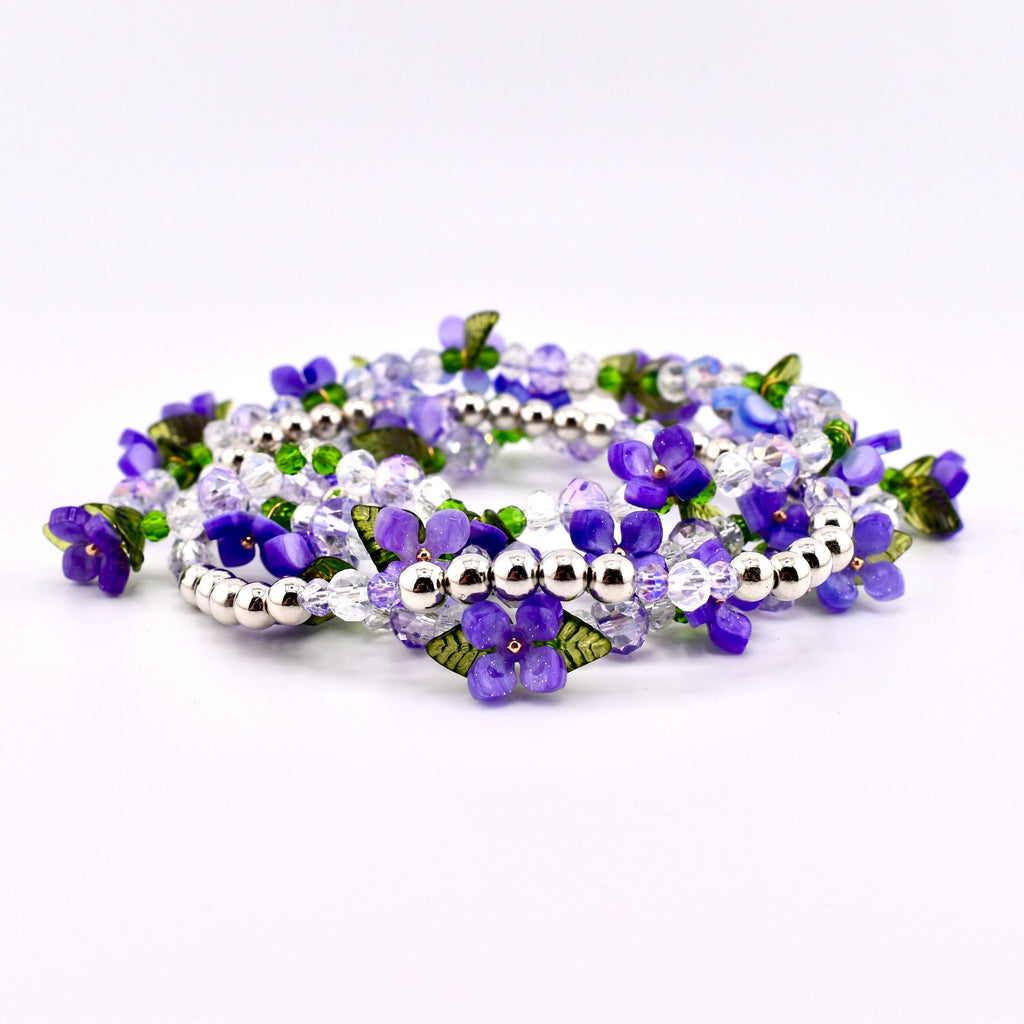 A collection of beaded bracelets featuring purple flower and leaf accents. The bracelets are made with clear and purple beads, silver-colored metal beads, and green leaf details. The bracelets are arranged in a circular pattern.