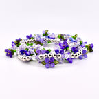 A collection of beaded bracelets featuring purple flower and leaf accents. The bracelets are made with clear and purple beads, silver-colored metal beads, and green leaf details. The bracelets are arranged in a circular pattern.