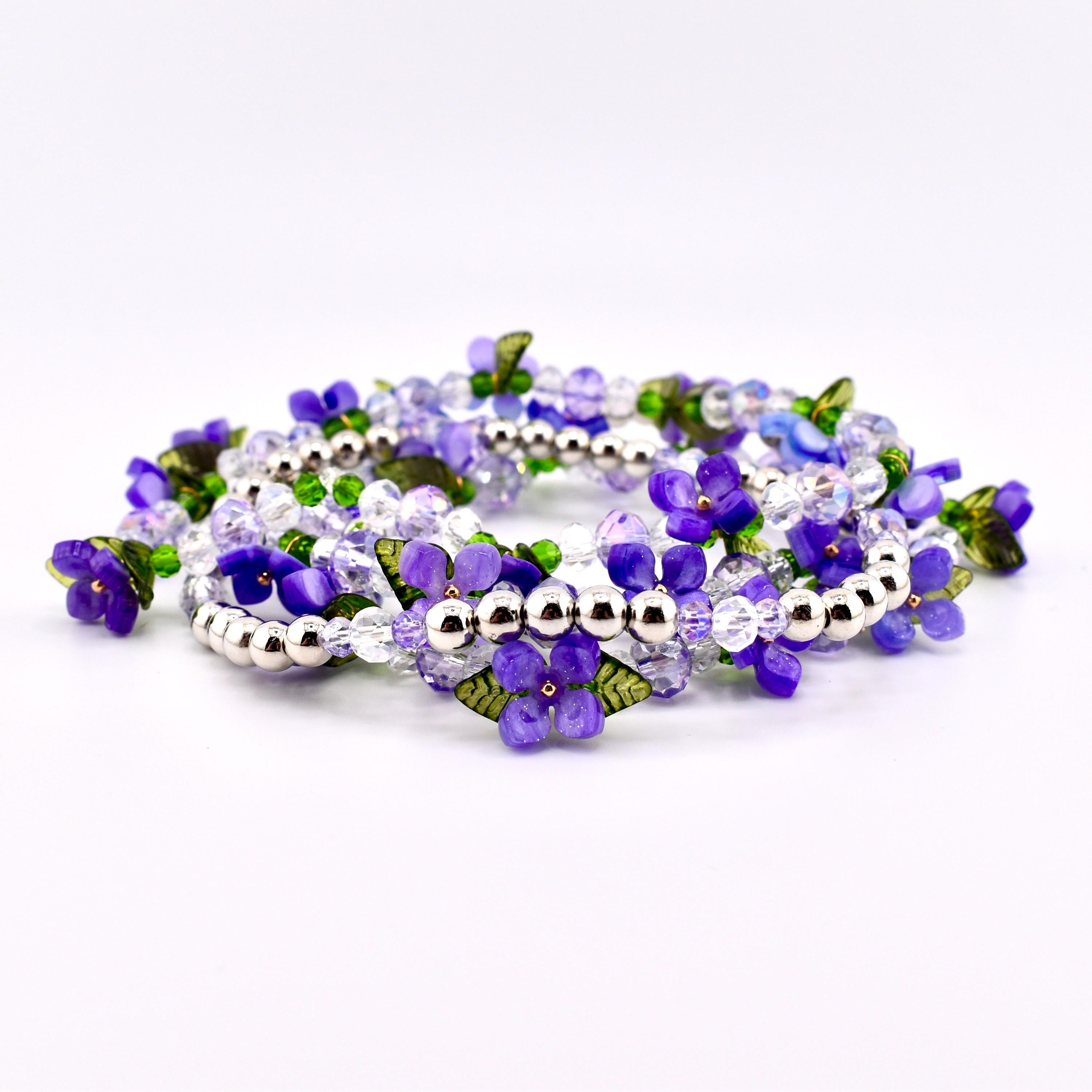 A collection of beaded bracelets featuring purple flower and leaf accents. The bracelets are made with clear and purple beads, silver-colored metal beads, and green leaf details. The bracelets are arranged in a circular pattern.